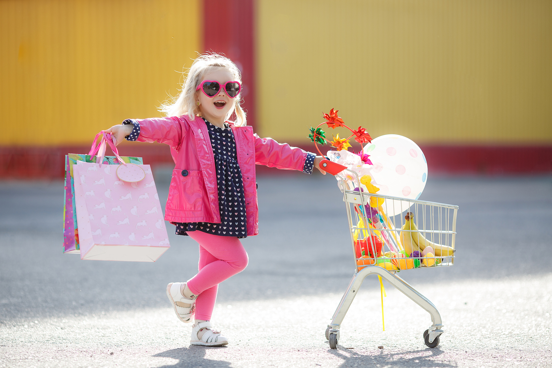 Girl three years old with a shopping trolley and paper bags in the parking lot near the supermarket.